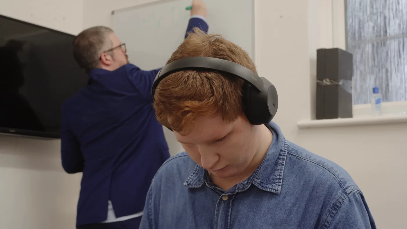 A student in headphones looks down at a desk while a teacher in a blue suit writes on a whiteboard behind him