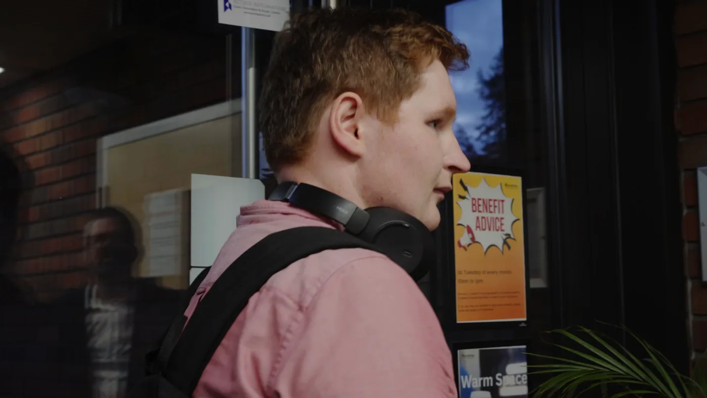 A young man in a pink shirt with headphones round his neck stands at a glass entrance beside a Benefit Advice poster